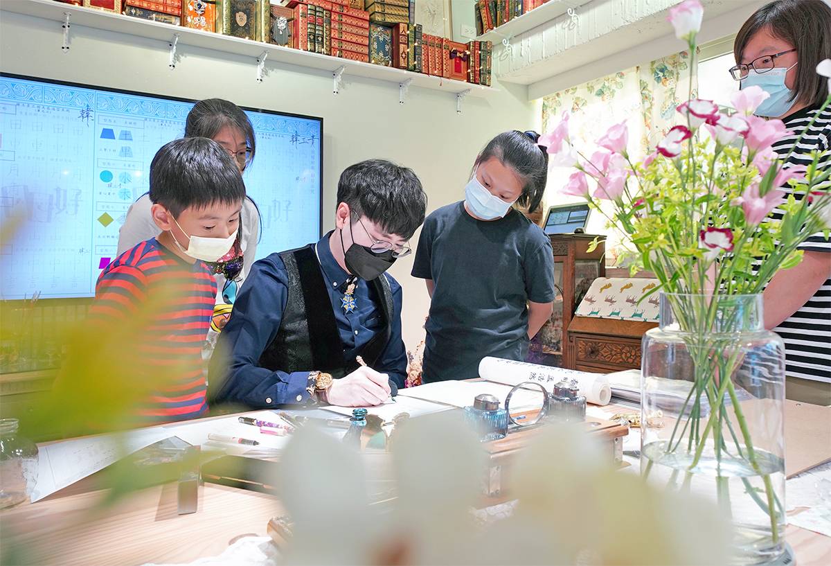 日日好文創老師於實體教室進行書寫示範，兒童與家長在旁近距離觀看，呈現親子共同參與藝術與文字學習的互動畫面。
